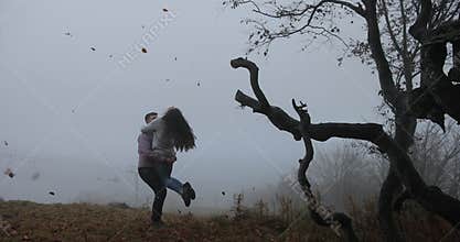 Dark autumn, mountain landscape. Man whirls woman before trees with fallen leaves covered with heavy fog