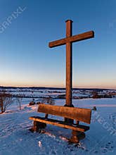 Wooden cross and bench on hill in snow-covered countryside by sunset light