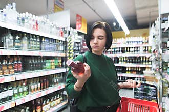 Portrait of a girl looking at a bottle of wine in her hand during shopping. Shopping for alcohol in a supermarket.
