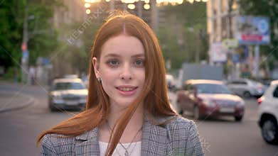 Young charming woman with ginger hair standing in street and looking in camera during sunset, smiling, traffic of cars