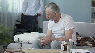 Nursing home. Handicapped man sitting on sofa. Male nurse bringing wheelchair