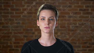Serious looking at camera caucasian woman with short hair and concentrated face, standing next to the brick wall chill