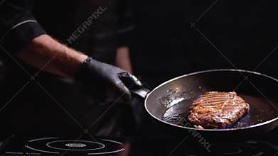 Male hand in black rubber gloves holding a frying pan with a grilling steak. The cook turns the pan on the fire in