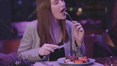 Beautiful young woman eating meat grilled with vegetable in the modern restaurant. The girl cuts one piece of meat and