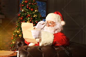 Portrait of happy Santa Claus sitting at his room at home near Christmas tree and reading Christmas letter or wish list.