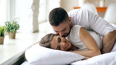 Young beautiful and loving couple wake up at the morning. Attractive man kiss and hug his wife in bed