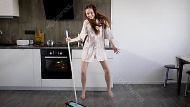 Funny girl is having fun, when doing house cleaning, dancing extravagantly around broom in kitchen