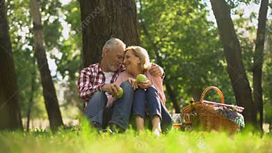 Joyful healthy old couple relaxing on grass, holding apples and hugging, picnic