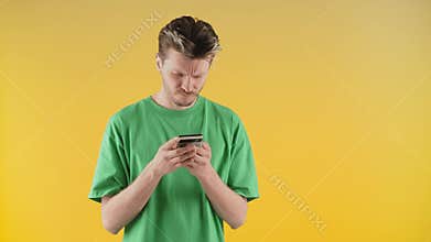 A young man in a green T-shirt stands with a phone in his hands and sighs heavily. Frustrated young man trying to