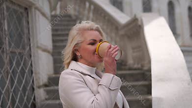A woman in a white coat is drinking coffee from a yellow cup. She is standing on a set of stairs.
