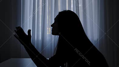 Silhouette of woman sitting at table in front of window, begging regretting expression, sad heartbroken mood, painful