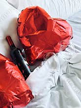 Two red heart-shaped balloons, two glasses and a bottle of red wine are on a white bed in a retro bedroom.