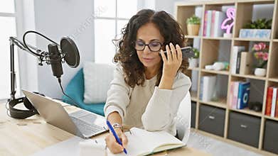 Hispanic woman podcasting indoor with microphone, laptop and notepad
