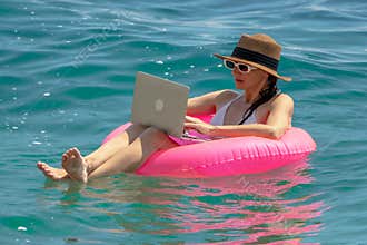 Remote work vacation freelancer woman typing on a laptop computer while floating in the turquoise blue ocean water.