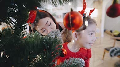 Child peeking through a Christmas tree adorned with ornaments during holiday preparations in a cozy home setting