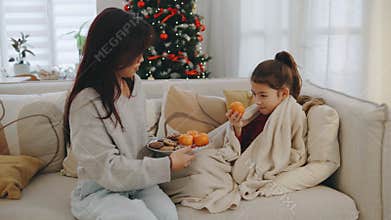 A mother shares fruits and cookies with her daughter, who is wrapped in a blanket, in a cozy living room decorated for the