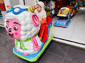 A young child wearing an orange outfit with a beige hijab is seated on a coin-operated ride shaped like a colorful cartoon sheep.