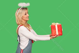 Happy birthday! Side view of angelic adult woman with halo above head giving gift box and smiling, offering present