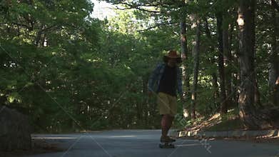 A young guy learns to ride a longboard. It is located in the forest. Dressed in a plaid shirt and a felt hat. Active