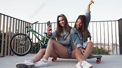 Two female best friends taking selfie with smartphone when sitting on skateboarding while walking at summer sunny day