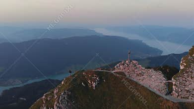 Aerial 4k orbital drone sunset at mountain peak with cross. Hiker girl climbing and taking pictures