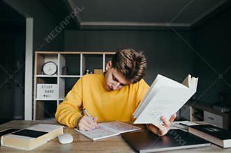 Young male student sits at a desk in a cozy room at home and studies, holding a book and writing in a notebook. Teenager is