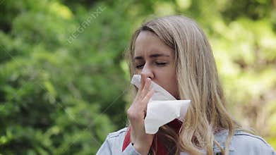 Young woman with with allergy symptom blowing nose standing in the park. Sick girl sneezing and blowing her nose into