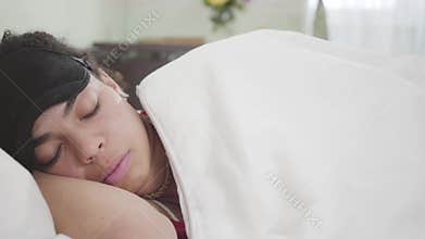 Close-up view of attractive African American woman lying under white blanket with black sleeping mask. Cute girl resting