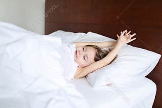 Little charming girl with raised hands sleeping on bed with white linens in morning.