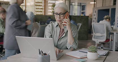 Confident mature business owner speaking on mobile phone and using laptop typing answering call in shared office