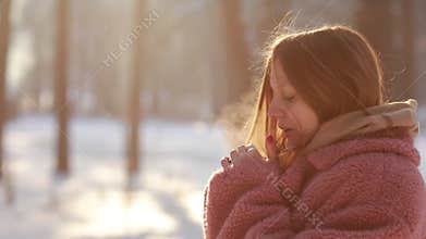 Beautiful young woman trying to warm her frozen hands with a breath in winter park. Girl with frosty breathing enjoying