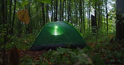 Green tent in forest with flashlight light inside. Unknown tourist in Carpathian mountains on rainy evening outdoors