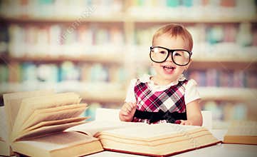 Funny baby girl in glasses reading book in library