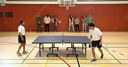 Playing table tennis, two men competing in school gymnasium with spectators watching