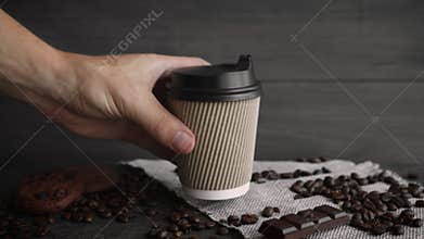 Hand placing disposable coffee cup on wooden table with roasted beans, chocolate, and cookies