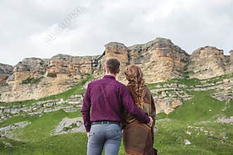 Portrait of a guy with a girl covered in a rug standing with his back in an embrace in nature and looking at the rocks.