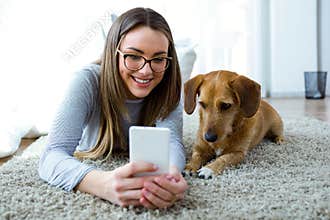 Beautiful young woman with her dog using mobile phone at home.