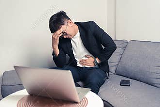 Young Asian business man sitting on sofa and working from home. Man feeling stress and suffering from headache and stomachache.