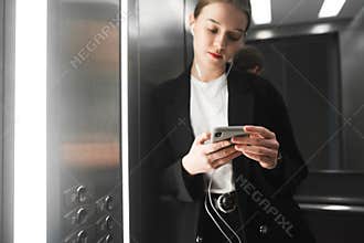 Close up photo of businesswoman holding smartphone in her heands in the elevator and listening to music in headphones. Portrait of