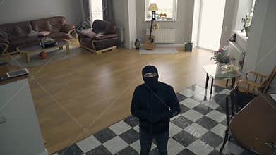 The thief in black clothes and balaclava walking in the large living room. Caucasian man raising hands noticing security