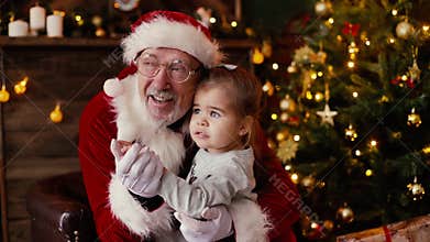 Little girl whispering Santa Claus ear her christmas wish sitting on his laps,