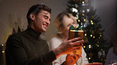 Side view of elegant mother-in-law giving festive gift box with Christmas present to happy man sitting with family at