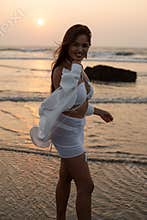 Carefree, laughing brunette female turn back to camera, wear white wet bikini, shirt, stand in sea water against sunset