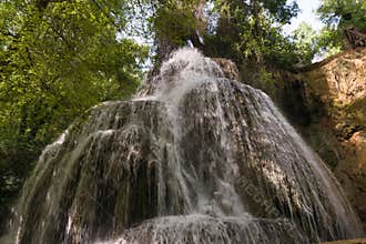 Waterfall cascades down rocky cliff surrounded by lush greenery in a tranquil natural setting on a sunny day