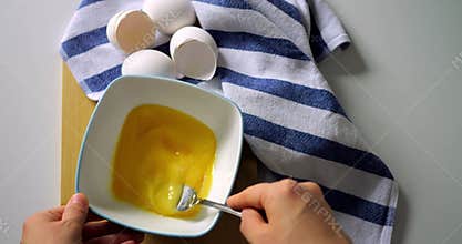Hand whisking beaten eggs in white bowl with fork on wooden table and striped towel, closeup top view of cooking