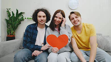 Three friends, a woman and two men, sit closely on a couch holding a large heart-shaped Valentine's Day card