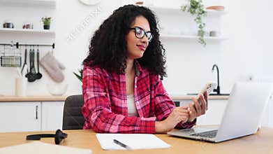 Young woman using smartphone at home while working remotely on a laptop