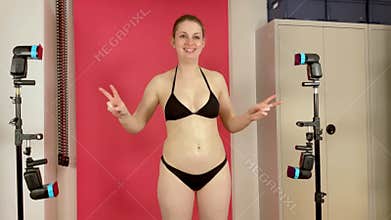 Pretty young woman in black bikini is posing in studio with pink background