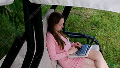 Young woman using laptop while sitting barefoot on garden swing outdoors working and relaxing in green countryside