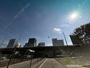 Sunny urban roadway beneath overpass with modern office towers, tram lines
